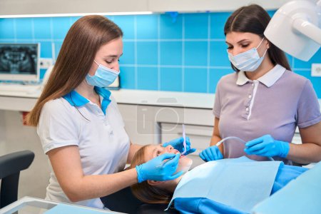 Foto de Asistente ayuda dentista en una recepción, ella sostiene un eyector de saliva, el médico trata un diente a un paciente joven - Imagen libre de derechos