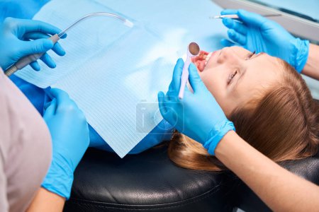 Téléchargez les photos : Adolescente dans la chaise des dentistes, le personnel médical utilise des instruments dentaires spéciaux au travail - en image libre de droit