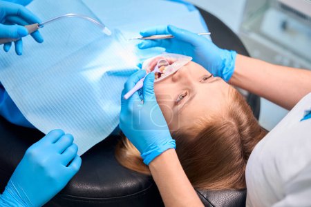 Téléchargez les photos : Le personnel médical de la clinique dentaire utilise des outils spéciaux pour nettoyer les dents, fille assise dans une chaise avec un rétracteur dentaire - en image libre de droit