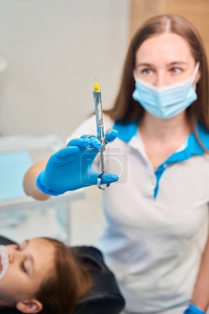 Female dentist holds syringe with local anesthesia in her hand, and teenage girl is sitting next to her in chair