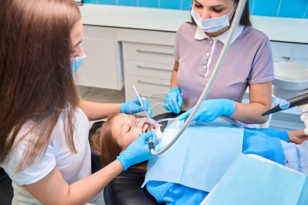 Foto de Empleado de una clínica dental perfora un diente de un paciente joven, un asistente tiene un eyector de saliva - Imagen libre de derechos