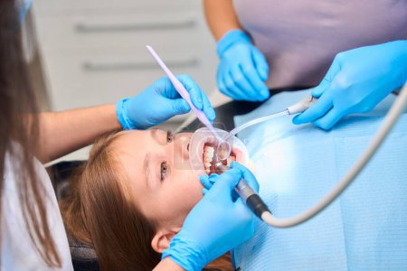 Female dentist drills a tooth of a teenage girl, an assistant holds a saliva ejector