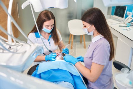 Female dentist with an assistant treats the teeth of a teenage girl, using modern equipment