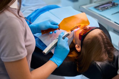 Doctor puts a photopolymer filling on the girl, the dentist uses a protective shield and glasses for the patient