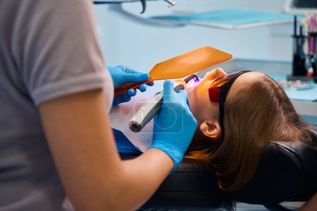 Dentist puts a photopolymer filling on the girl, she uses a protective screen and glasses for the patient