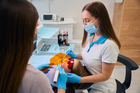 Dentist puts a photopolymer filling on a girl, an assistant helps her