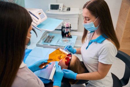 Femme dentiste met un photopolymère remplissage sur une fille, un assistant l'aide