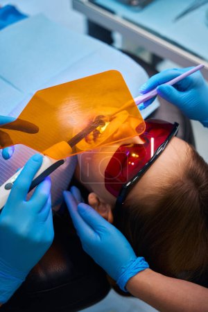 Médecin dentiste utilise une lampe photopolymère moderne et un écran de protection au travail, une jeune patiente porte des lunettes de sécurité