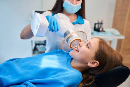 Téléchargez les photos : Docteur tient une lampe photopolymère dentaire dans ses mains, il voit une adolescente à son rendez-vous - en image libre de droit