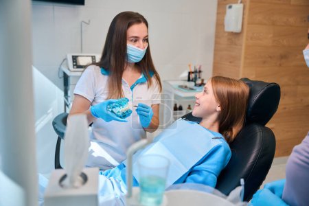 Female hygienist communicates during a consultation with a young patient, she has a model of the dentition in her hands