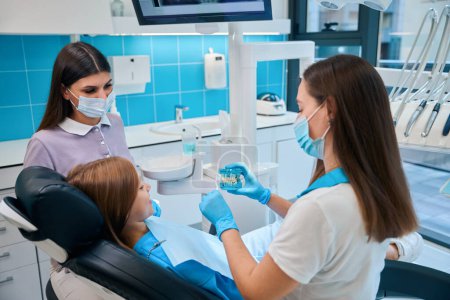 Foto de La higienista femenina demuestra un modelo de dentición a una paciente joven, con un joven asistente cerca - Imagen libre de derechos