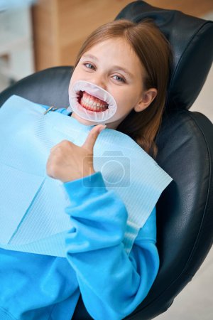 Foto de Chica adolescente se sienta en una silla dental y se muestra bien, en el retractor dental - Imagen libre de derechos