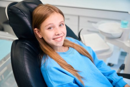 Smiling girl sitting in a dental chair, wearing a blue sweatshirt