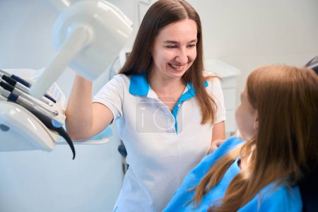Téléchargez les photos : Hygiéniste dentaire communique gentiment avec une jeune patiente dans un cabinet dentaire, dans une pièce avec un équipement moderne - en image libre de droit