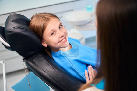 Foto de Sonriente adolescente se comunica con una higienista femenina en un consultorio dental, en el interior con equipos modernos - Imagen libre de derechos