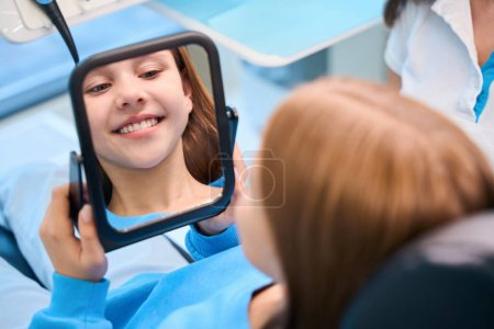 Foto de Joven paciente admira su sonrisa de dientes blancos en el espejo, la chica está en el consultorio dental - Imagen libre de derechos