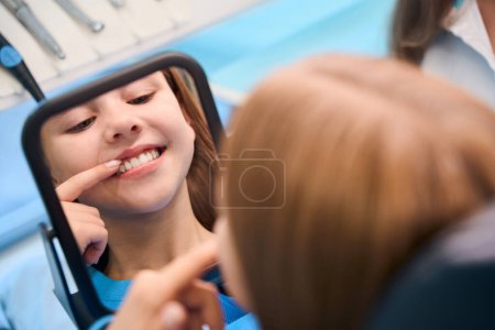 Téléchargez les photos : Fille admire son sourire à dents blanches dans le miroir, elle est dans un cabinet dentaire - en image libre de droit