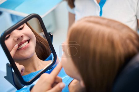 Foto de Niño admira su sonrisa de dientes blancos en el espejo, ella está en el consultorio de dentistas - Imagen libre de derechos