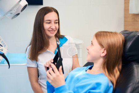 Foto de Chica en un consultorio dental mira sus dientes en el espejo, al lado de una mujer dentista - Imagen libre de derechos