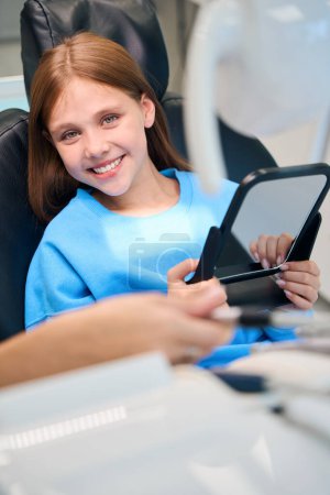 Foto de Adolescente en recepción en una clínica dental, se sienta en una silla dental con un espejo en las manos - Imagen libre de derechos