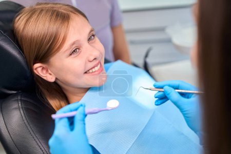 Foto de Chica sonriente se sienta en la silla de dentistas, el especialista comienza a examinar los dientes de las niñas - Imagen libre de derechos
