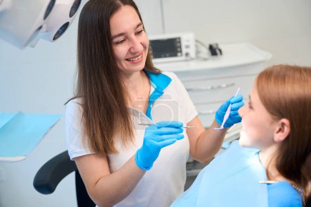 Foto de Dentista joven comienza a examinar la cavidad oral de un paciente joven, la sudadera de las niñas está cubierta con una servilleta azul - Imagen libre de derechos