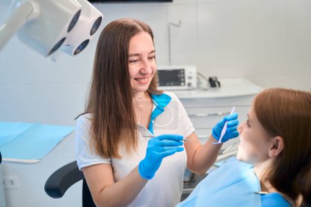 Foto de Higienista femenina comienza a examinar la cavidad oral de un paciente joven, la sudadera de las niñas está cubierta con una servilleta azul - Imagen libre de derechos
