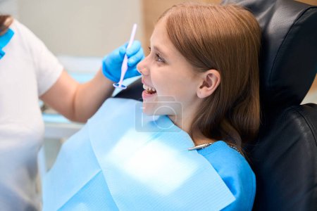 Téléchargez les photos : Adolescente souriante communique avec un dentiste au bureau, le médecin a un miroir dentaire dans ses mains - en image libre de droit