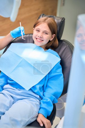 Foto de Especialista comienza a examinar la cavidad oral de un paciente joven, la sudadera de las niñas está cubierta con una servilleta azul - Imagen libre de derechos