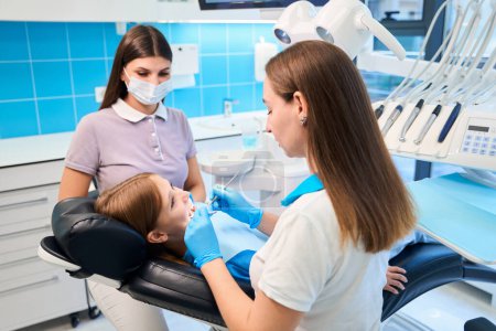 Foto de Higienista dental examina los dientes de una niña, junto a ella es una asistente femenina en una máscara protectora - Imagen libre de derechos