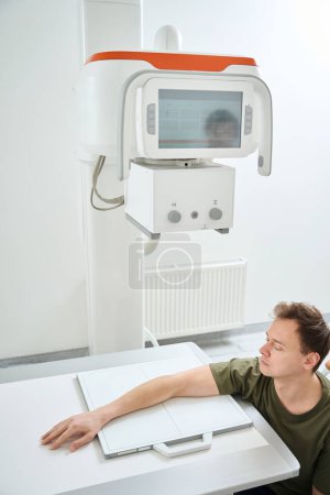 Male person sitting at end of radiography table with his arm placed on image receptor