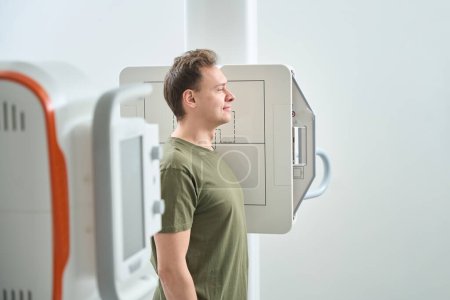 Photo for Side view of man standing upright with his left arm positioned against image receptor in front of radiography machine - Royalty Free Image