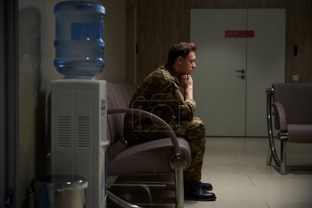 Foto de Vista lateral del hombre militar reflexivo que descansa la barbilla en las manos apretadas mientras está sentado en el sofá cerca del dispensador de agua en el pasillo - Imagen libre de derechos