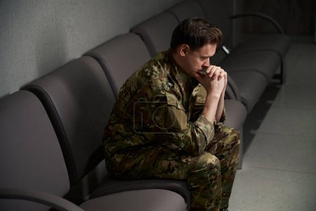 Dejected serviceman in camouflage uniform propping his chin with thumbs seated on leather sofa in waiting area