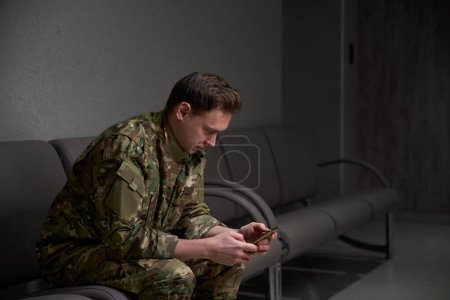 Serious focused military man in camouflage uniform looking at smartphone screen while sitting on leather sofa in building corridor