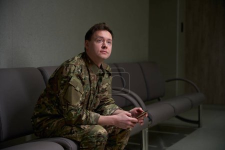 Ruhiger Soldat in Tarnuniform mit Handy in der Hand auf dem Sofa im Wartebereich
