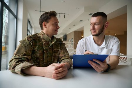 Smiling medical professional making notes while sitting at reception desk in waiting room beside pleased military man