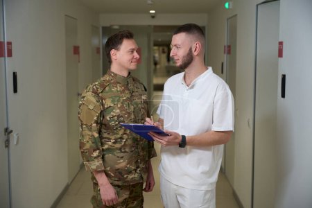 Focused doctor writing on clipboard while looking at pleased serviceman in clinic corridor