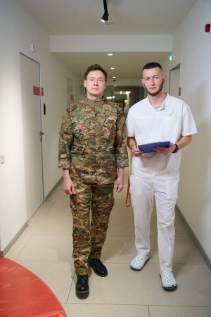 Full-size portrait of physician with clipboard and pen in hands standing beside serviceman in medical facility hallway