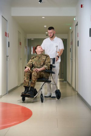Ganztägiges Porträt des Gesundheitshelfers, der Rollstuhl mit Militärmann durch den Flur der medizinischen Einrichtung schiebt