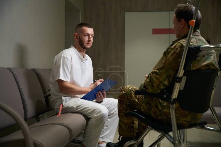 Arzt macht sich Notizen auf Klemmbrett, während er den im Rollstuhl sitzenden Soldaten in der Halle der medizinischen Einrichtung betrachtet