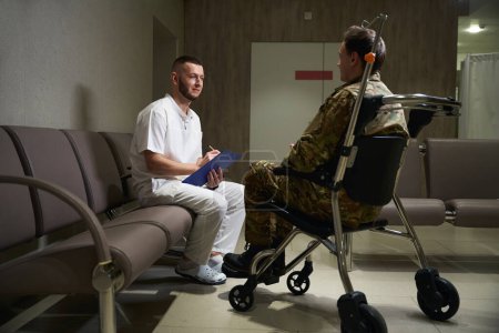 Foto de Médico con lápiz y portapapeles en las manos mirando al militar sentado en silla de ruedas en el pasillo del hospital - Imagen libre de derechos