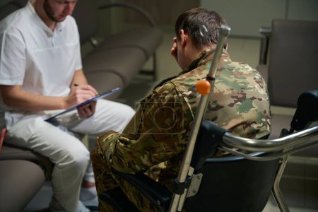 Ernsthafter Gesundheitsexperte macht sich Notizen mit Stift auf Klemmbrett in Anwesenheit eines Soldaten im Rollstuhl im Krankenhausflur