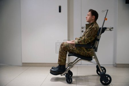 Photo for Side view of military man seated in wheelchair in healthcare facility hallway looking into distance - Royalty Free Image