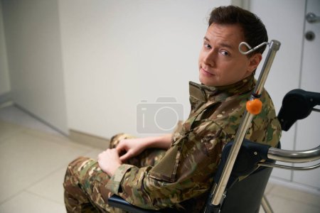 Trauriger Soldat sitzt im Rollstuhl im Flur der medizinischen Einrichtung und blickt in die Kamera