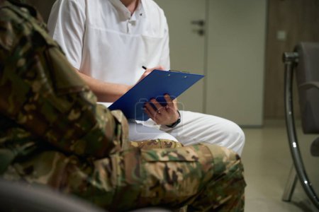 Cropped photo of doctor making notes while sitting on sofa next to military man in hospital hall