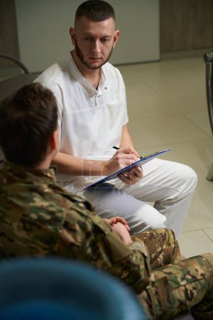 Mediziner sitzt im Krankenhausflur und schaut den Soldaten an, während er Notizen mit Stift auf Klemmbrett macht