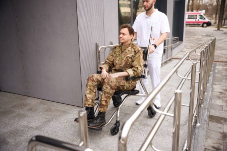 Téléchargez les photos : Infirmière masculine sérieuse poussant fauteuil roulant avec jeune militaire vers le bâtiment de l'hôpital - en image libre de droit