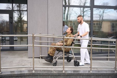 Foto de Vista lateral del trabajador médico empujando la silla de ruedas con el militar hacia la puerta principal del hospital - Imagen libre de derechos