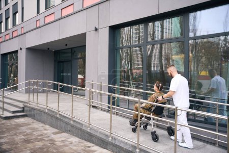 Nurse pushing wheelchair with disabled person in camouflage uniform towards healthcare facility front door